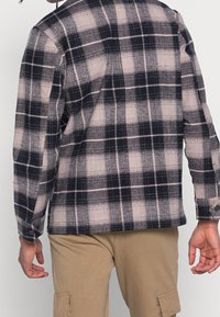 Košile s dlouhým rukávem v kostkovaném vzoru černé, bílé a béžové barvy. Má volný střih, zapínání na knoflíky na manžetách a rovný spodní lem s měkkou texturou.