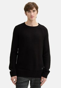 Schwarzer Strickpullover mit langen Ärmeln, gerippten Bündchen und Saum, strukturiertem Stoff und Rundhalsausschnitt, mit einem kleinen Etikett-Detail am Saum.