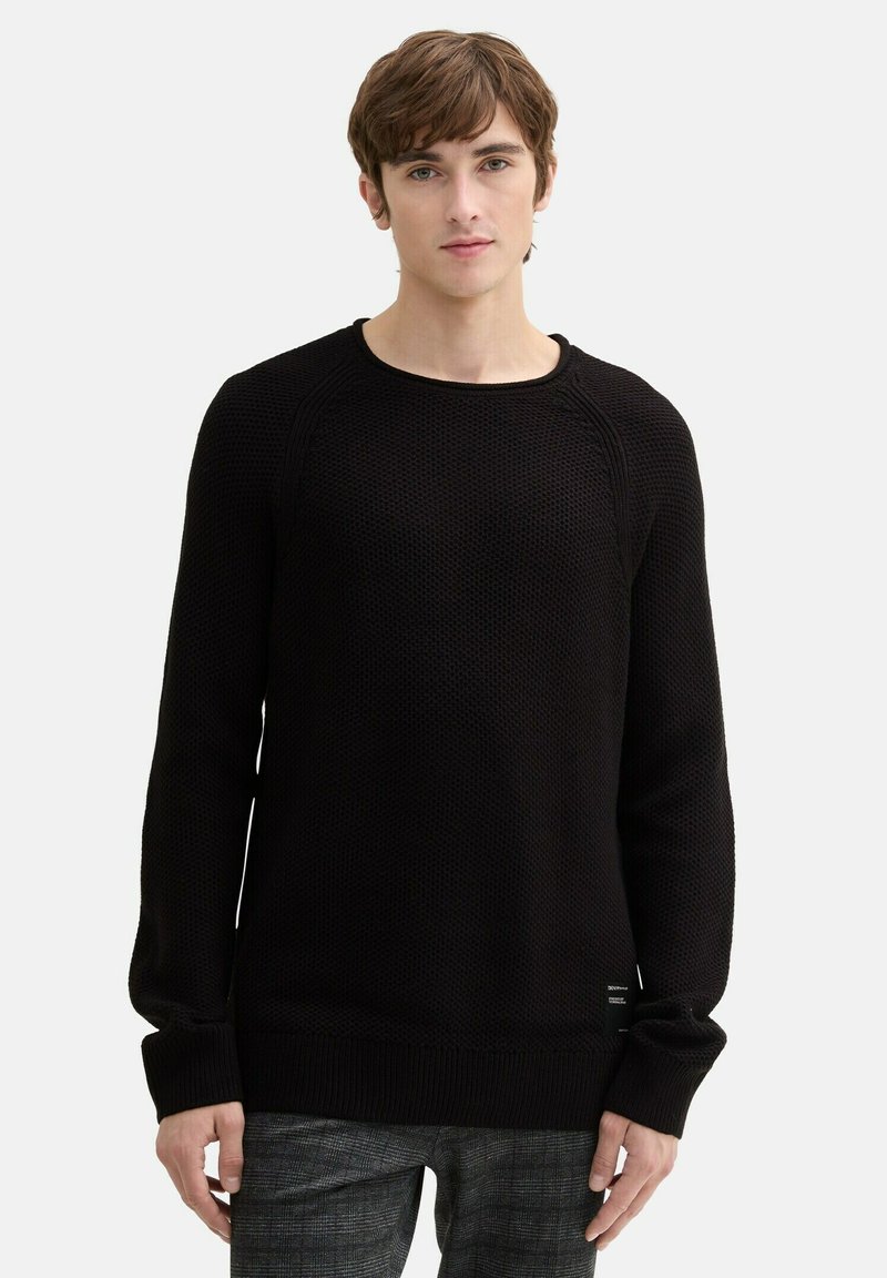 Schwarzer Strickpullover mit langen Ärmeln, gerippten Bündchen und Saum, strukturiertem Stoff und Rundhalsausschnitt, mit einem kleinen Etikett-Detail am Saum.