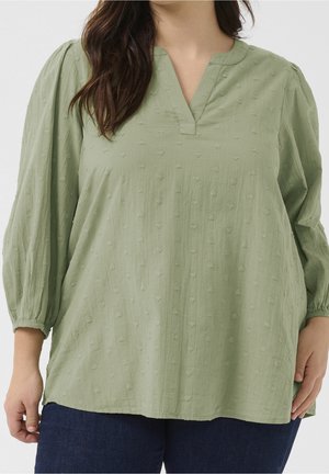 Woman wearing a light green textured blouse with three-quarter puff sleeves and a V-notch neckline, paired with dark blue pants.