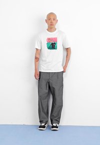Hvit bomulls t-skjorte med et fargerikt grafikktrykk på forsiden, sammen med grå oversized bukser og svarte joggesko.