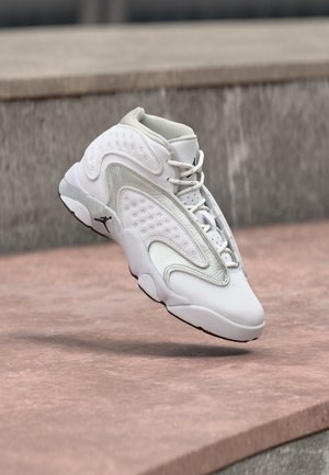 Λευκό sneaker ψηλού τύπου με πολύπλοκο σχεδιασμό πάνελ και μαύρη ενδιάμεση σόλα με λογότυπο, εμφανίζεται στον αέρα πάνω από μια πέτρινη επιφάνεια.