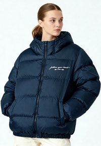 Veste matelassée bleu marine avec capuche, design matelassé et poignets élastiques. Comprend la phrase brodée "suis ton cœur" en blanc.