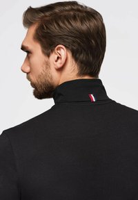 Schwarzes Poloshirt mit hohem Kragen, ausgestattet mit einem kleinen dreifarbigen Akzent (rot, weiß, blau) am Hals. Glatte Textur, schlichtes Design.