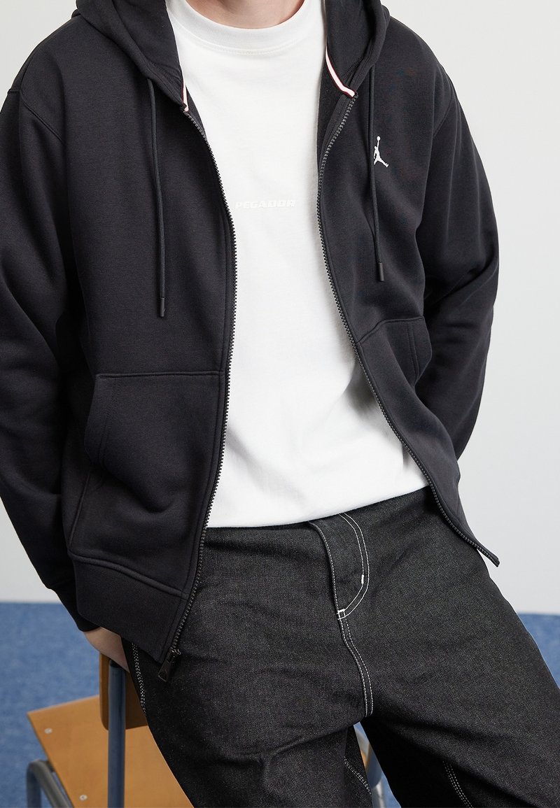 Persona che indossa una felpa nera con zip e logo bianco, una t-shirt bianca e jeans scuri, seduta su una sedia di legno davanti a uno sfondo semplice.