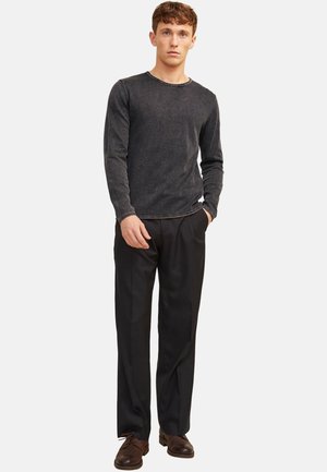 Junger Mann, der ein ausgewaschenes schwarzes Langarmshirt, schwarze Hose und braune Lederschuhe trägt, mit einer Hand in der Tasche.