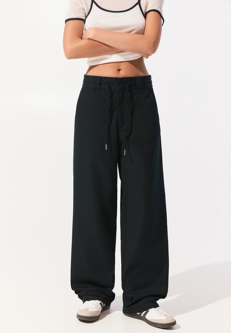 Black wide-leg pants feature a drawstring waist, smooth fabric, and subtle sheen. Paired with white sneakers and a fitted top.