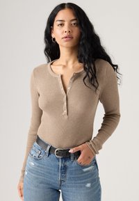 Top beige de manga larga con cuello redondo y botones en la parte frontal, combinado con unos jeans de mezclilla azul desgastados y un cinturón negro.