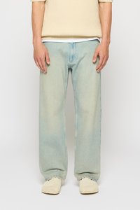 Persoană îmbrăcată cu un pulover bej deschis, jeans lejeri de culoare albastru deschis și pantofi slip-on bej stând pe un fundal alb simplu.