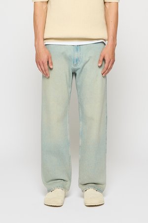 Person in einem hellbeigen Pullover, lockeren hellblauen Jeans und beigen Slippern, der vor einem schlichten weißen Hintergrund steht.
