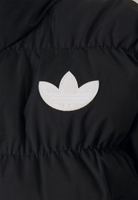 White Adidas trefoil logo patch stitched on black quilted fabric, likely a jacket or coat.