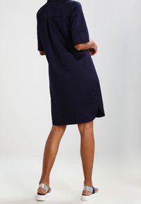 Mörkblå skjortklänning i bomullsblandning, med korta ärmar, krage och sidosten. Bärs med ljusa avslappnade sandaler.