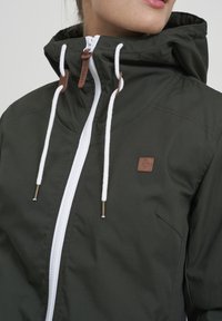Dark green jacket with a high collar, white zipper, and drawstrings. Features brown leather accents and a side pocket.
