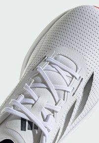 Zapatilla deportiva blanca con parte superior de malla, detalles texturizados y cordones delgados. Presenta acentos en negro y una suela roja.