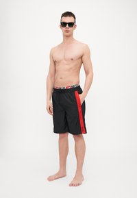 Black swim shorts with a red side stripe, elastic waistband featuring logo, and drawstring, made from lightweight material.
