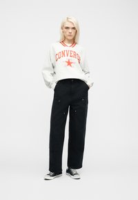 Ξανθός/ή άτομο στέκεται με τα χέρια στις τσέπες, φορά γκρι φούτερ Converse, μαύρο παντελόνι με φαρδιά μπατζάκια και μαύρα χαμηλά αθλητικά παπούτσια.