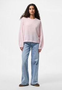 Woman with long curly hair wearing a light pink sweater, light blue wide-leg jeans, and brown shoes standing against a white background.