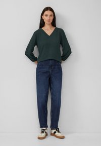 Donkergroen, gestructureerd V-hals top met losse mouwen, gecombineerd met hooggeplaatste blauwe spijkerbroeken en witte sneakers met zwarte accenten en een bruine zool.