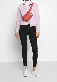 Chaqueta de mezclilla rosa claro con bolsillos, jeans negros ajustados y zapatillas blancas. Una bolsa cruzada roja descansa sobre el pecho.