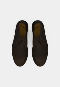 Chaussures en daim brun foncé avec un bout rond, des lacets bicolores et une surface texturée. L'intérieur présente une semelle intérieure de couleur claire contrastante.