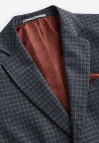 Šedý kostkovaný blazer se špičatými klopy, který má bohatou podšívku v barvě rezavé a kapesníček v kontrastním odstínu. Texturovaná tkanina.
