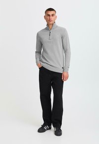 Grå zip-up tröja med långa ärmar, ribbat nederkant och mudd, parat med svarta byxor och svarta sneakers, står mot en enkel bakgrund.