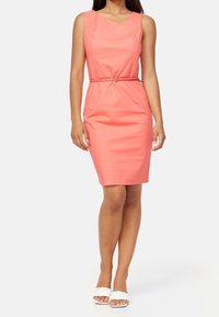 Korálové přiléhavé šaty s čtvercovým výstřihem a tenkými ramínky, stažené v pase kulatým opaskem. Doplněné o bílé sandály.