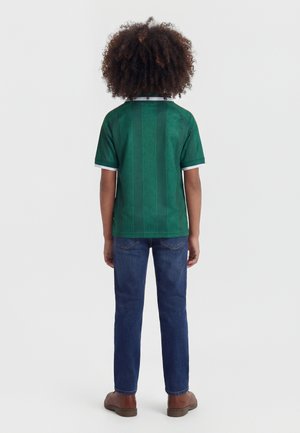 Enfant aux cheveux bouclés, de dos, portant un t-shirt vert à manches courtes, un jean bleu et des chaussures marron sur fond blanc.