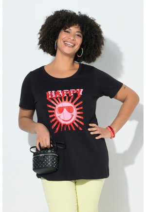 Lächelnde Frau mit lockigem Haar trägt schwarzes T-Shirt mit dem Schriftzug „HAPPY“ und rotem Sonnenmotiv, gelbe Hose, Regenbogen-Hoop-Ohrringe, hält kleine schwarze Tasche mit Nieten.