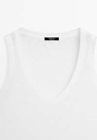 Top blanc sans manches avec col en V et texture en maille fine, comportant une étiquette noire portant l'inscription "Massimo Dutti Made in China" à l'intérieur du col.