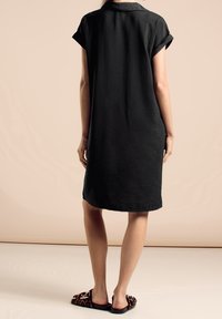 Schwarzes, knielanges Hemdkleid mit weichem Textil und umgeschlagenen Ärmeln. Kombiniert mit flachen Sandalen im Leopardenmuster.