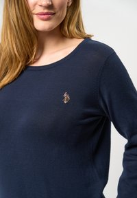 Temno modra, dolga majica srokav, s kroženjm ovratnikom, izdelana iz mehkega materiala; ima majhen izvezen logotip na prsih.