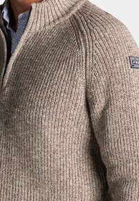 Beige gebreide trui met een hoge ribbelkraag, voorzien van gestructureerde patronen en een merklabel op de schouder.