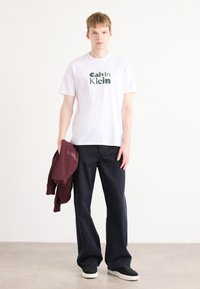 Hvit t-skjorte med grønn "Calvin Klein" logo, mørkeblå vide jeans og en burgunder hettegenser som henger over armen. Sorte sneakers på føttene.