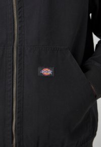 Juoda zip-up striukė pagaminta iš patvarios medžiagos. Turi ryškų kišenę, gumuotus rankogalius ir matomą prekės ženklo emblemą ant kišenės.