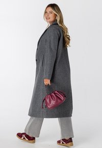 Vrouw die loopt, gekleed in een lange grijze jas, brede grijze broeken, bordeauxrode sneakers en met een bijpassende bordeauxrode handtas.