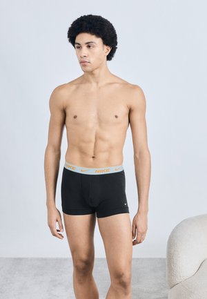 Čierne boxerky Nike s svetlosivým pásom s oranžovým nápisom. Model pózuje na jednoduchom pozadí. Hladká textúra látky.