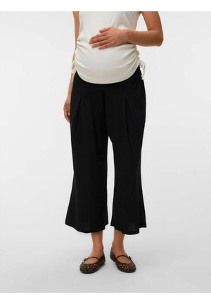 Gravid person iført en hvid rynket top, sorte brede cropped bukser og brune vævede flats, hånd hvilende på maven.
