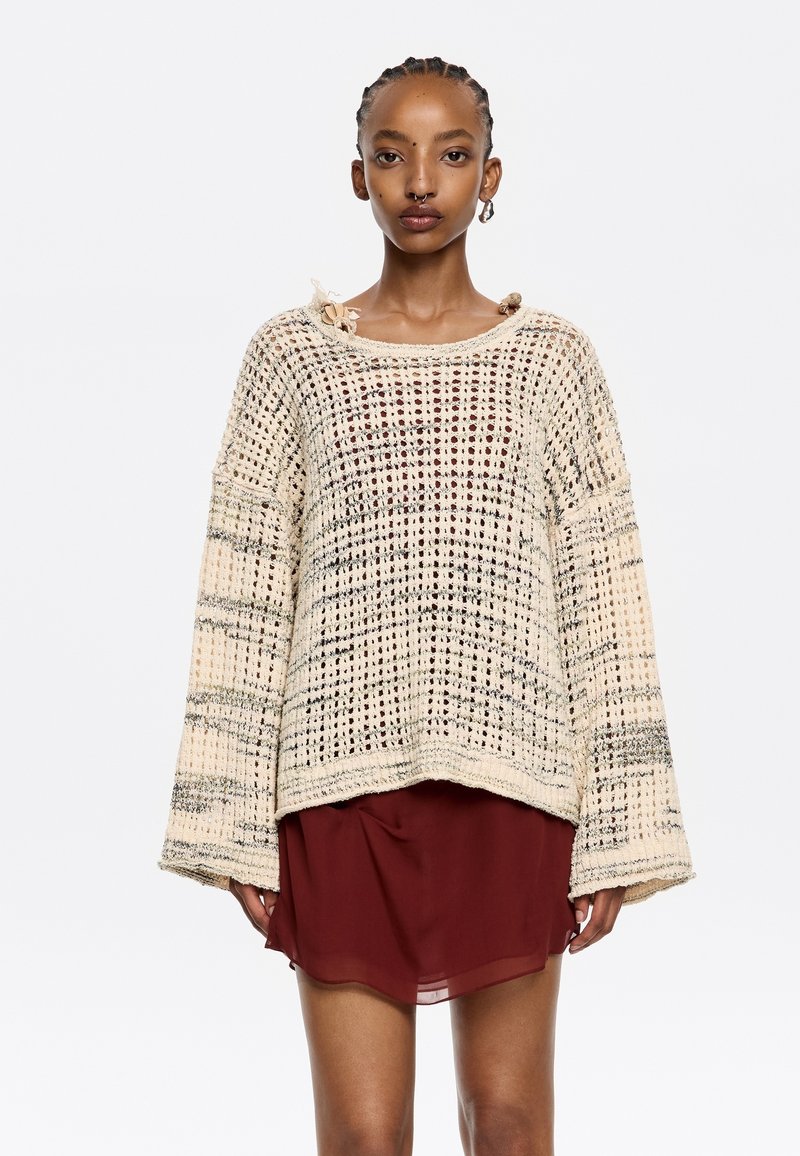 Strickpullover in Creme mit einer lockeren, übergroßen Passform, der ein strukturiertes Muster und weite Ärmel aufweist. Kombiniert mit einem kurzen burgunderfarbenen Kleid.