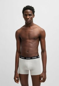 Graue Baumwoll-Boxershorts mit schwarzem Bund, der das Logo "HUGO" zeigt. Das Modell steht mit freiem Oberkörper vor einem neutralen Hintergrund.