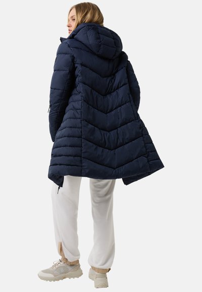 Marineblå pufferjakke med chevron-syninger, hætte og lynlåslommer. Båret over hvide bukser og sneakers, der viser en afslappet, casual pasform.