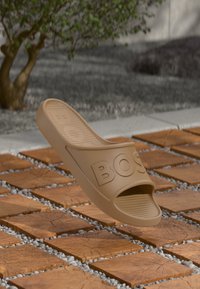 Sandale à glissière en caoutchouc marron clair avec une large bride ornée du logo "BOSS" en relief. Design à bout ouvert et semelle texturée, surélevée au-dessus d'une surface en carreaux à motifs.