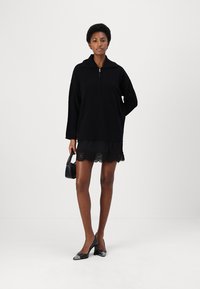 Felpa nera zipata sovradimensionata con maniche lunghe, indossata sopra una gonna nera con bordo in pizzo. Modella che tiene una piccola borsa nera e indossa tacchi neri.