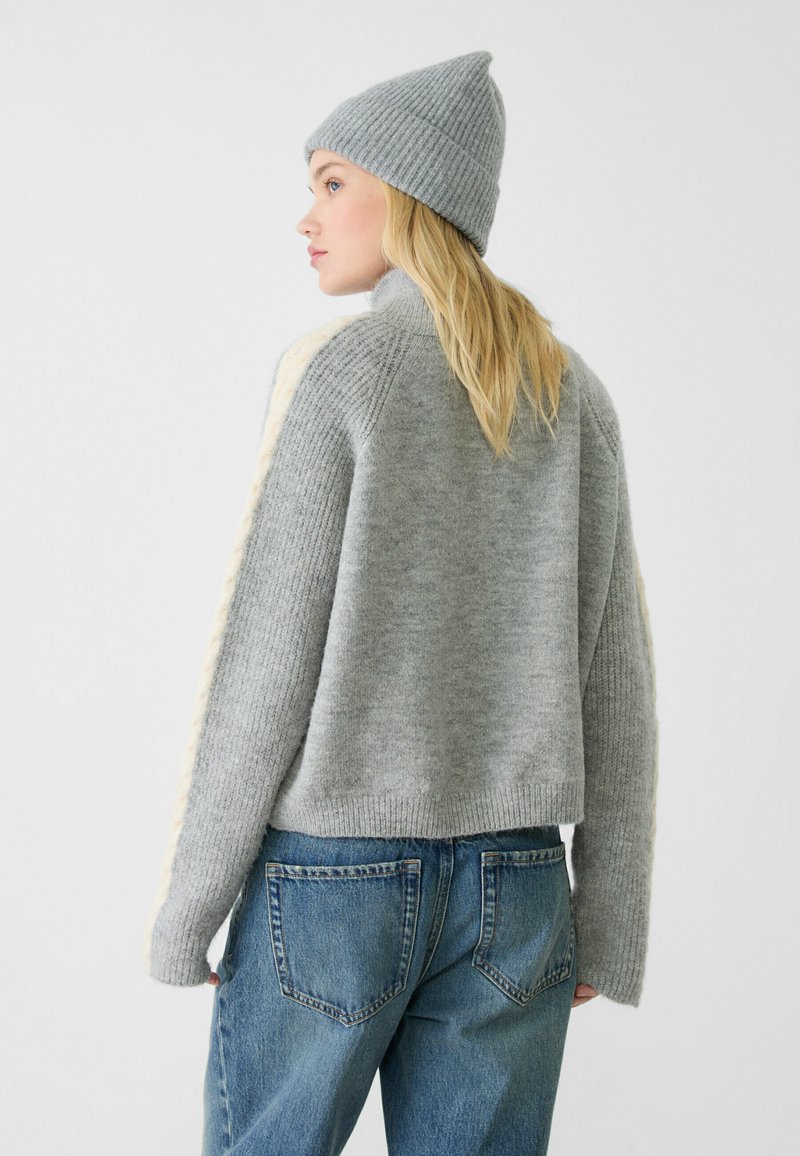 Pullover grigio a maglia con texture a coste e accenti crema, abbinato a jeans in denim e un berretto grigio coordinato.
