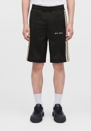 Mand iført sorte shorts i midt-længde med beige sidelstriber, hvide sokker, sorte sneakers og en hvid skjorte mod en ensfarvet baggrund.