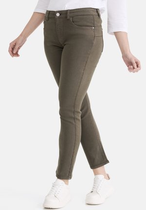 Olivgrüne Denim Skinny Jeans mit mittlerer Bundhöhe, versehen mit abgenähten Nähten, vorderen Taschen und einem Reißverschluss-Detail, kombiniert mit weißen Turnschuhen.