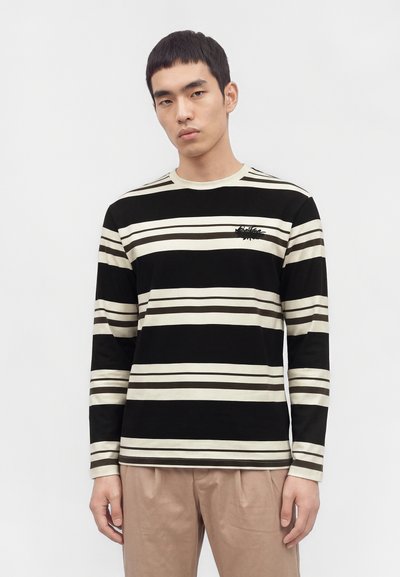 Jeune homme portant un t-shirt à manches longues rayé horizontalement en noir et blanc et un pantalon beige, debout devant un fond clair uni.