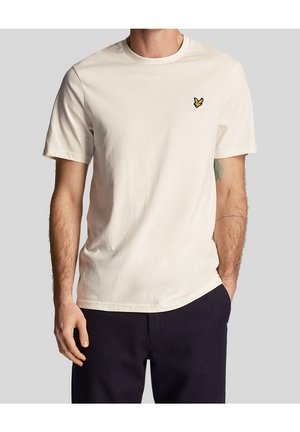 Homme portant un t-shirt uni blanc cassé à manches courtes avec un petit logo d'oiseau jaune sur la poitrine, associé à un pantalon foncé sur un fond uni.