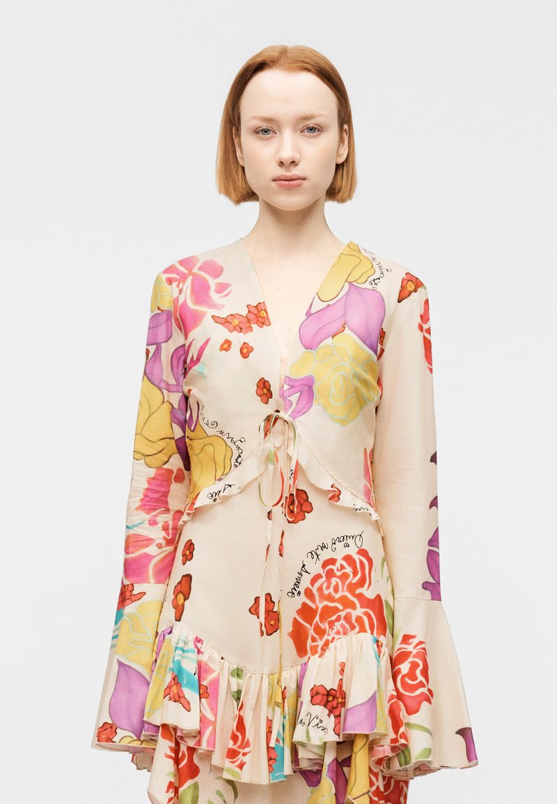 Νεαρή γυναίκα με ίσια κόκκινα μαλλιά που φοράει μπεζ φόρεμα με λουλούδια σε μοβ, κίτρινο και κόκκινο χρώμα, με βολάν στο τελείωμα και στα μανίκια.