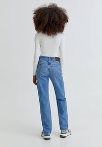 Jeans blu chiaro con vestibilità dritta, vita alta e tasche posteriori. Indossati con una maglietta bianca a maniche lunghe e sneakers grigie.